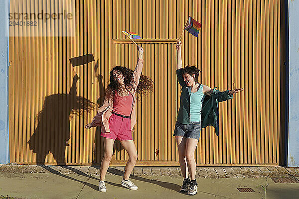 Excited lesbian couple dancing and raising LGBTQIA pride flag in front of brown building wall