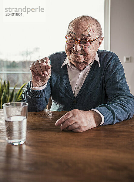 Retired senior man holding medicine sitting at table with glass of water