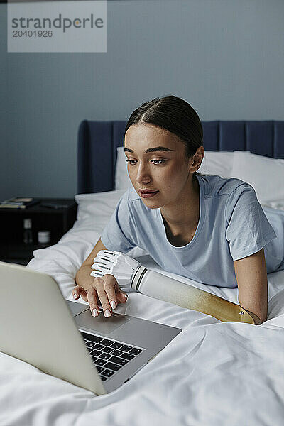 Young woman with arm prosthesis using laptop lying on bed in bedroom