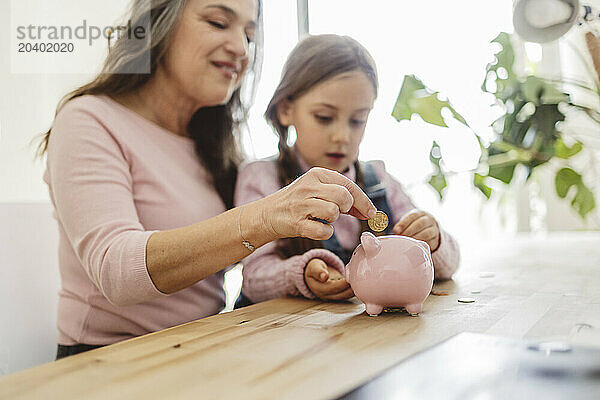 Grandmother and granddaughter inserting coin into piggy bank at home