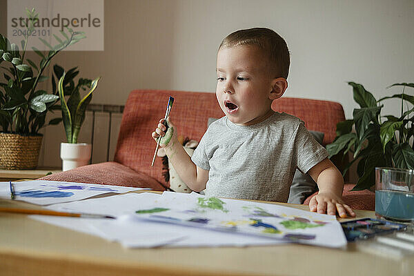 Surprised boy holding paintbrush with papers on table at home