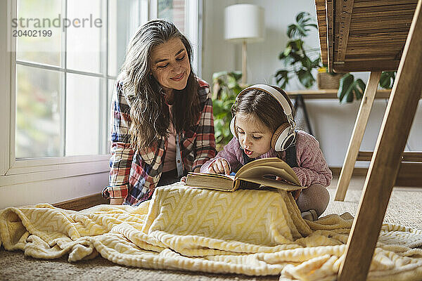 Granddaughter listening to music and reading book sitting by smiling grandmother at home