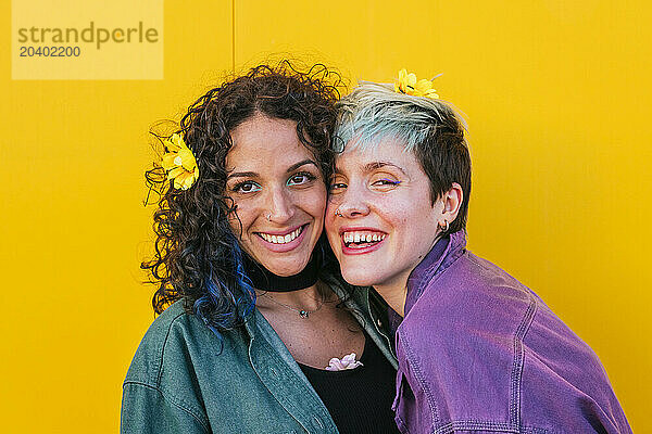 Cheerful lesbian couple with flowers on hair in front of yellow wall