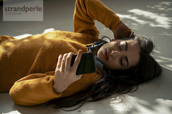 Woman with hand in hair using smart phone lying on floor at home