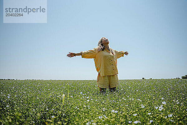 Woman wearing yellow linen clothing standing with arms outstretched in blooming field on sunny day