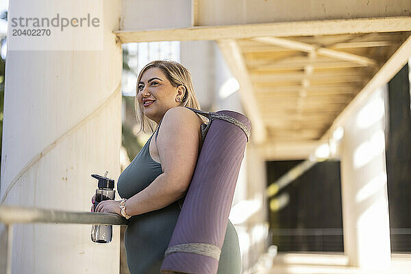 Smiling young curvy woman standing with exercise mat and water bottle