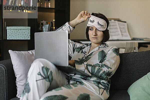 Young woman in pajamas and eye mask looking at laptop working from home