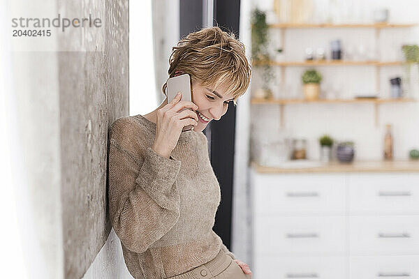 Smiling woman talking through smart phone near wall