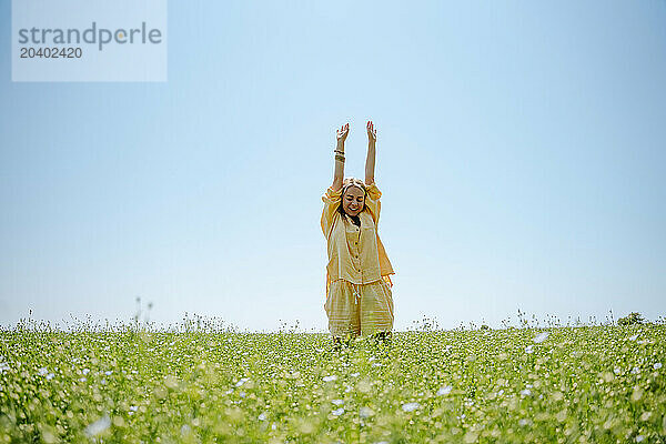 Woman wearing yellow linen clothing standing with arms raised in blooming field on sunny day