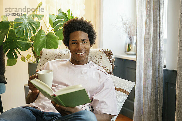 Smiling man with coffee cup reading book reclining on lounge chair at home