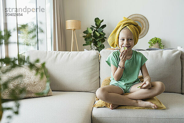 Smiling girl sitting on sofa and eating cucumber at home