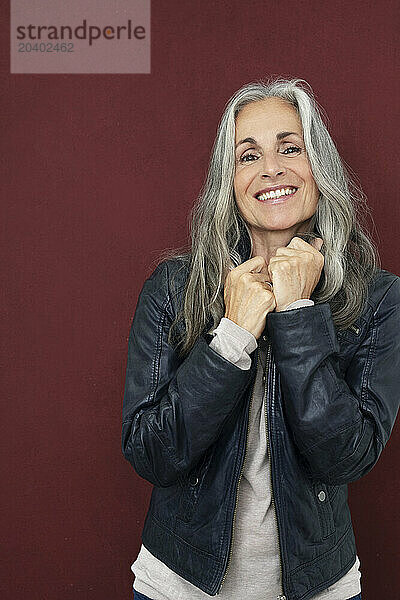 Smiling mature woman in leather jacket standing against maroon background