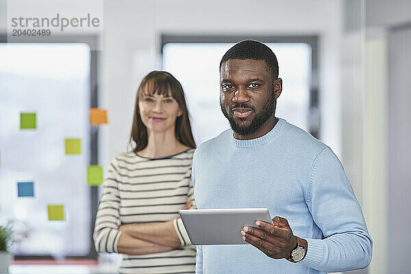 Confident businessman holding tablet PC with coworker in background