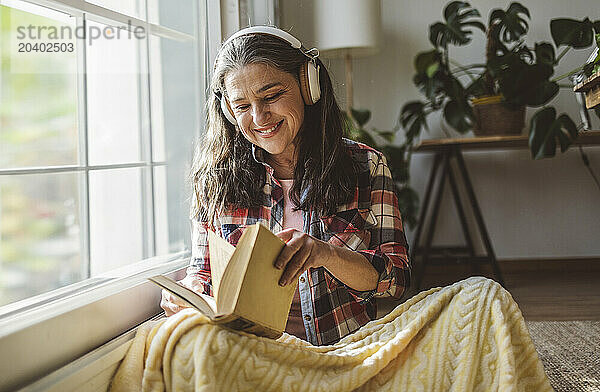 Smiling mature woman listening to music through wireless headphones reading book at home