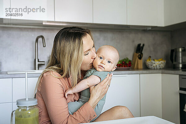 Blond woman kissing toddler son in kitchen at home