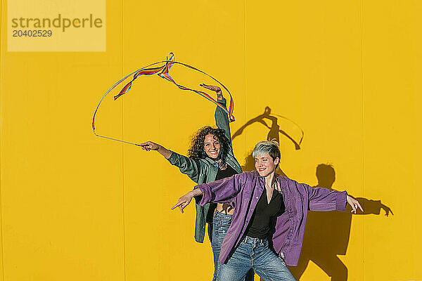 Smiling woman waving multi colored ribbon with lesbian friend in front of yellow wall