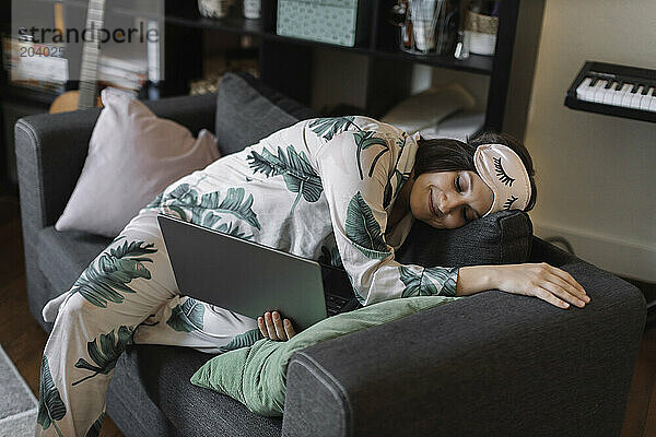 Tired businesswoman in pajamas and eye mask sleeping with laptop on sofa home