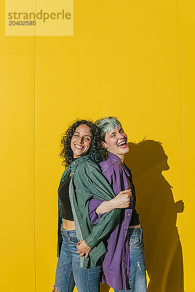 Happy lesbian couple standing back to back in front of yellow wall