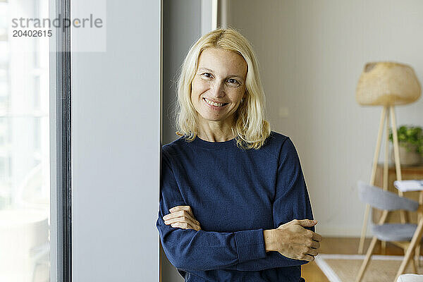 Smiling mature woman leaning with arms crossed on window at home