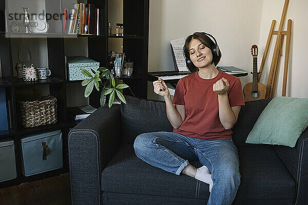 Smiling woman with eyes closed listening to music through wireless headphones sitting on sofa at home