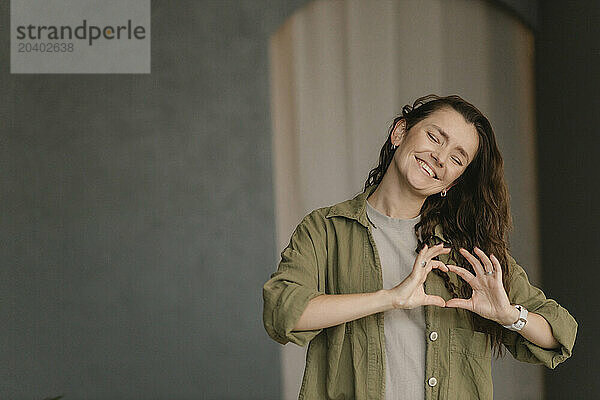 Happy woman with eyes closed making heart shape standing in new home