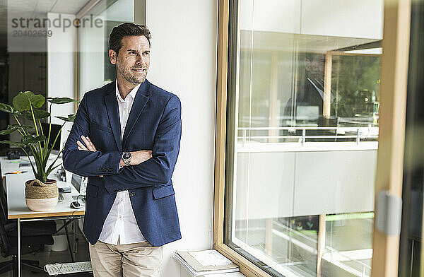 Businessman standing with arms crossed looking out through window of office