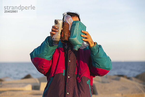 Man in jacket holding plastic bottles in front of face near breakwater