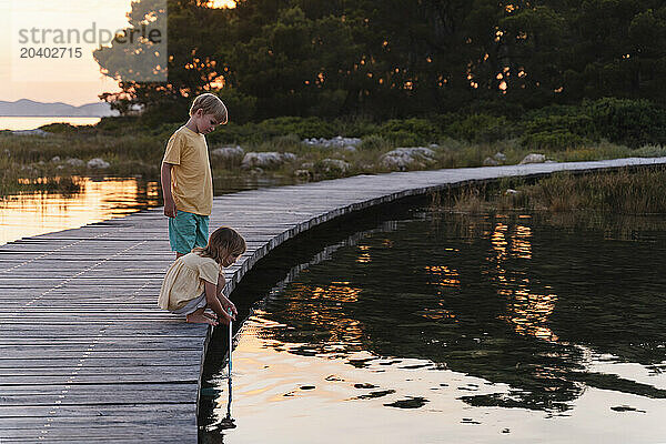 Girl crouching with fishing net by brother standing in lake at sunrise