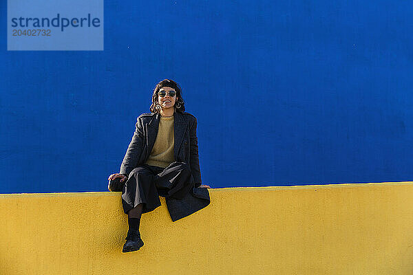 Smiling woman sitting on top of yellow wall