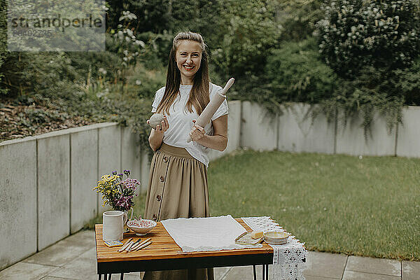 Smiling woman holding clay and rolling pin standing by table at back yard