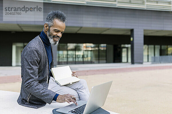 Smiling mature businessman working on laptop and sitting with diary near building