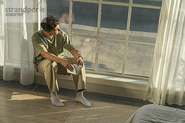 Thoughtful young man sitting with book near window