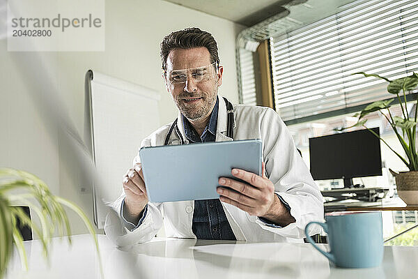 Smiling doctor wearing smart glasses using tablet PC sitting at desk in office