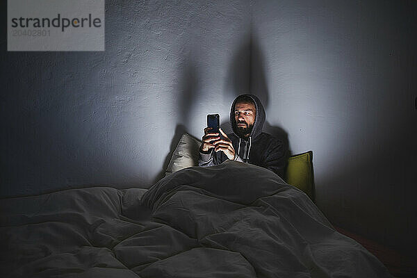 Man using smart phone sitting on bed in dark room