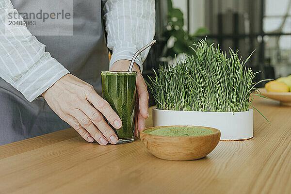 Woman holding glass of wheatgrass juice in kitchen at home