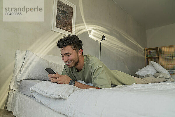 Happy young man lying on bed and using smart phone at home