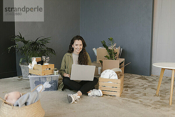 Smiling woman using laptop sitting amidst boxes in new home