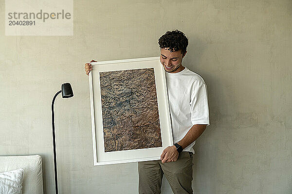 Happy man standing with picture frame near wall at home