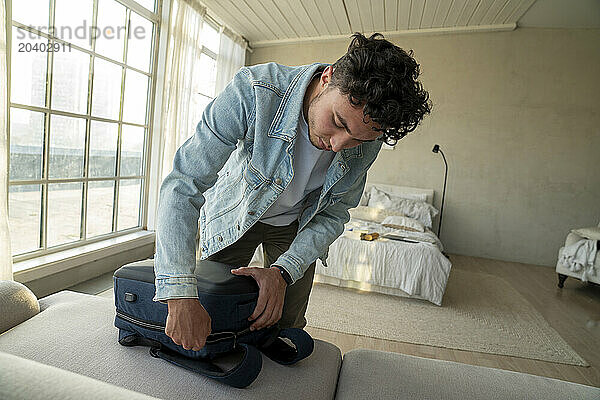 Young man closing zip on backpack on sofa at home