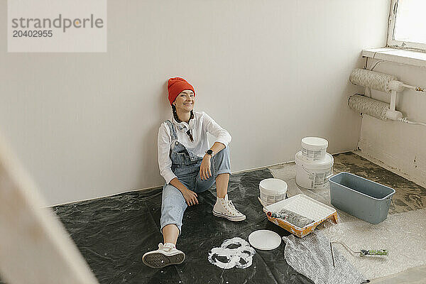 Smiling woman leaning on white wall sitting by paint equipment in new apartment