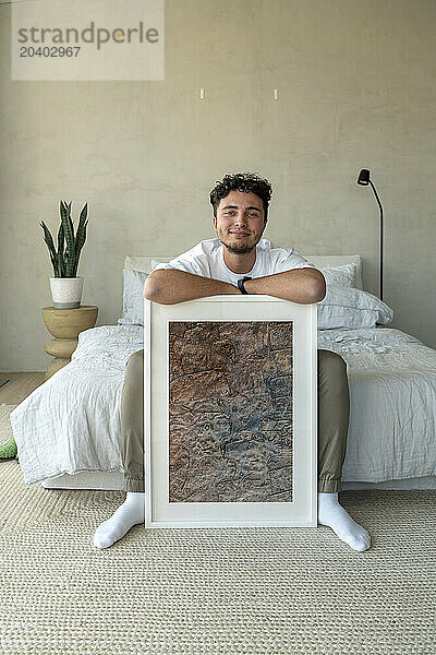 Smiling young man sitting with picture frame on bed at home