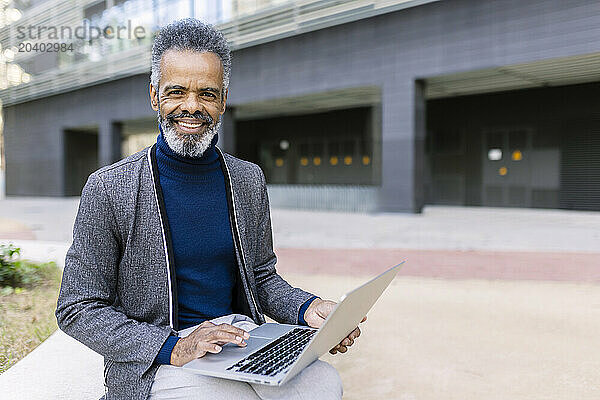 Happy businessman sitting with laptop in front of building