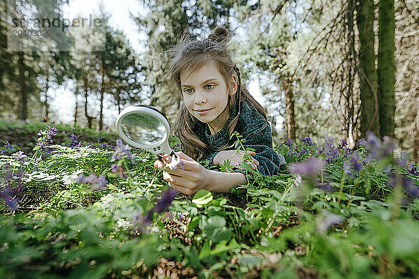 Smiling girl looking at flowers with magnifying glass in forest