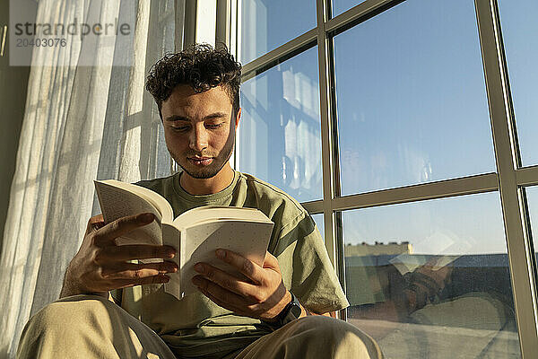 Young man reading book near window at home