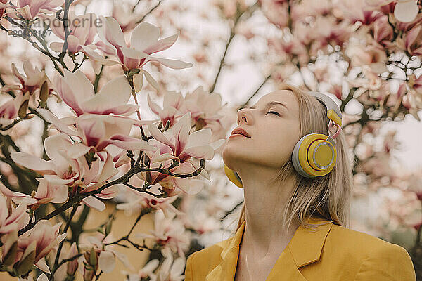 Blond woman listening to music through wireless headphones with eyes closed by magnolia tree in garden