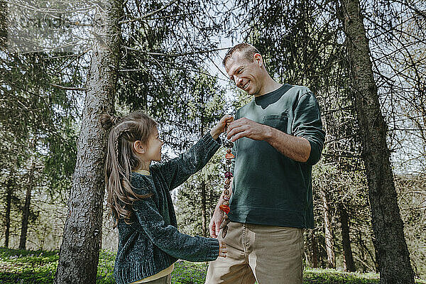 Smiling father and daughter holding natural necklace standing near trees in forest