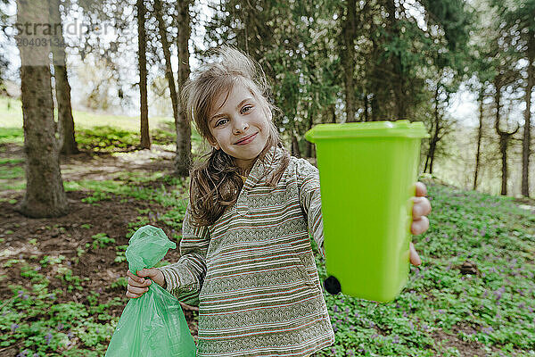 Smiling girl holding plastic bag and showing garbage bin toy at forest