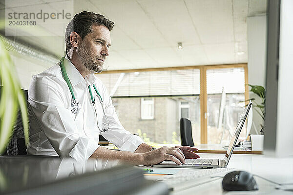 Mature doctor using laptop sitting at desk in office