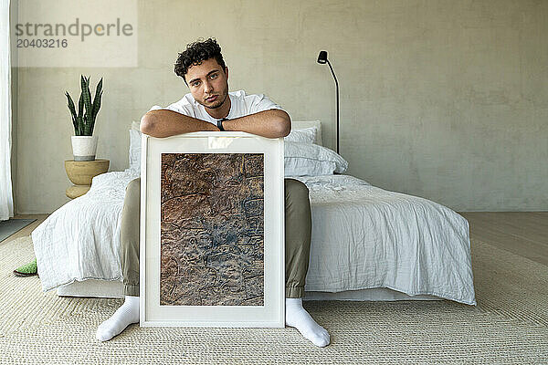 Young man sitting with picture frame on bed at home