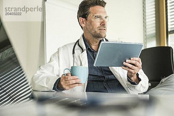 Doctor wearing smart glasses using tablet PC sitting at office desk in clinic